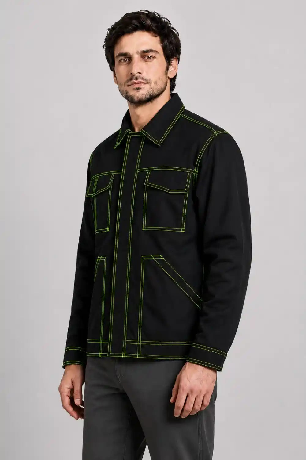 A man wearing a black utility-style jacket with bright green contrast stitching and gray trousers, photographed against a plain light gray studio background.