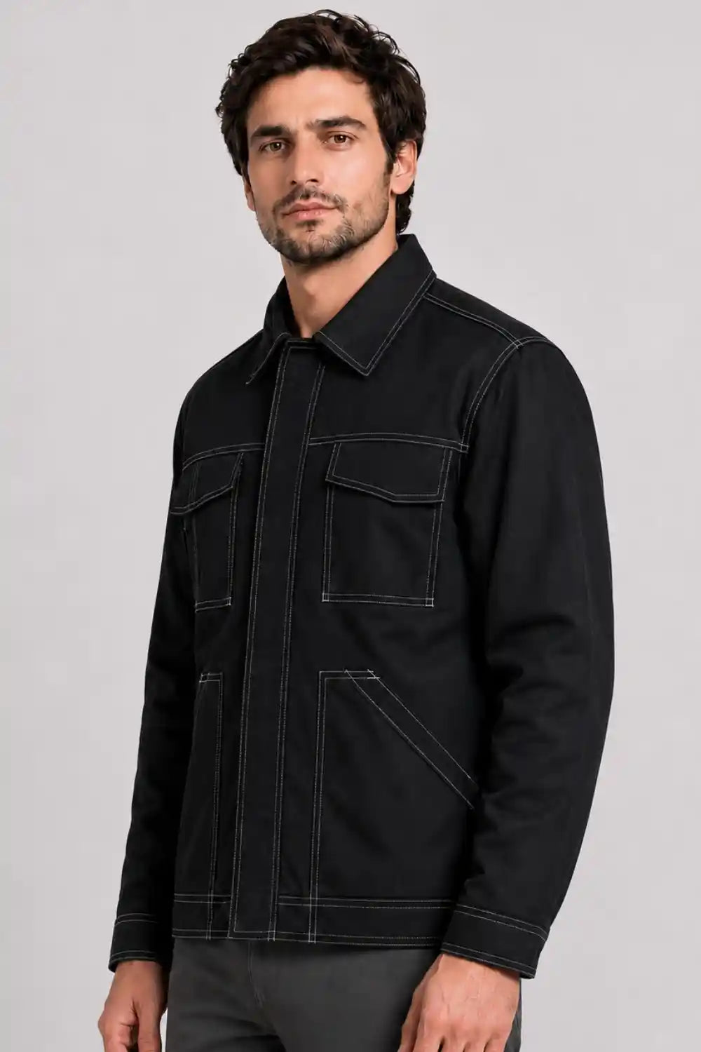 A man wearing a black utility-style jacket with contrasting white stitching, photographed against a neutral light gray studio background.