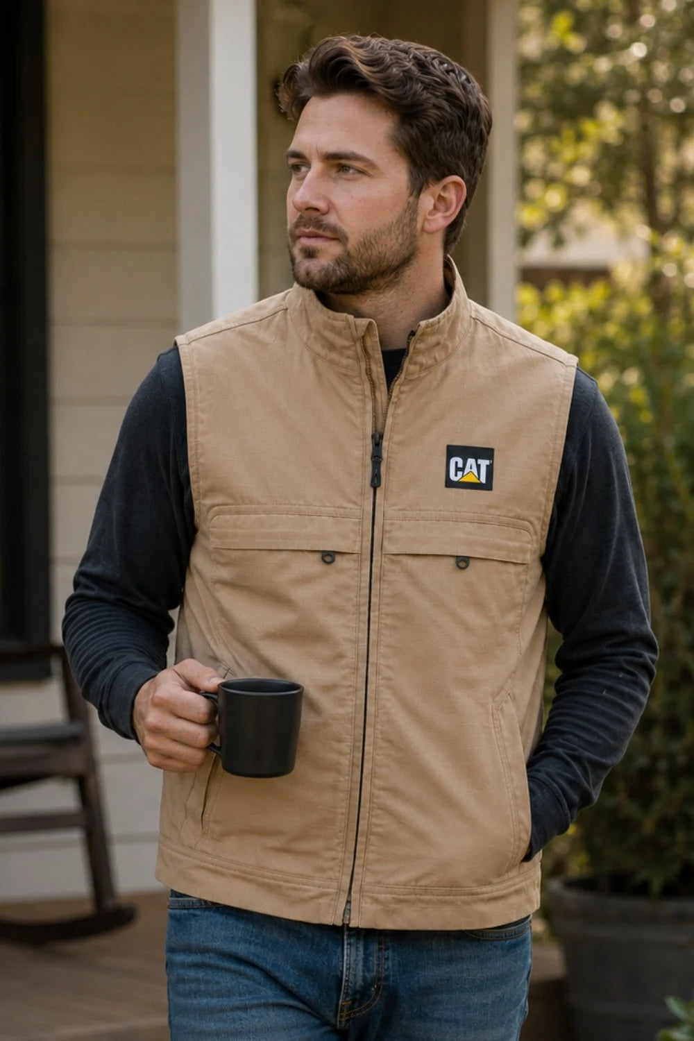 Man wearing a tan vest with a Cat logo, holding a black mug outdoors.